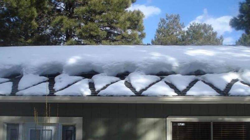 Snow on a roof forms triangular patterns across the shingles, with trees and a blue sky in the background.