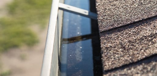 Close-up view of a house roof with asphalt shingles and a rain gutter filled with standing water. Close-up view of a house roof with asphalt shingles and a rain gutter filled with standing water.