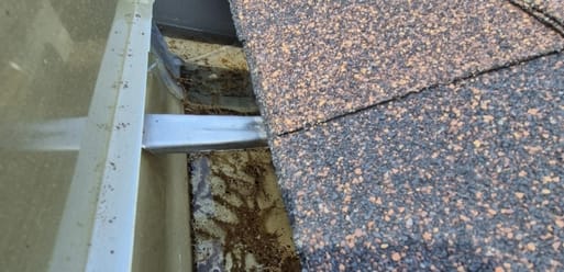 Close-up view of a rooftop with shingle tiles, exposed flashing, a metal support bar, and debris collected in the gutter area. Close-up view of a rooftop with shingle tiles, exposed flashing, a metal support bar, and debris collected in the gutter area.