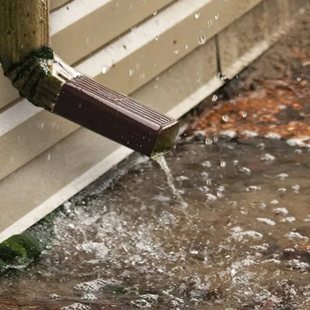Water flows from a brown downspout onto the ground, causing splashing and pooling near a building's exterior wall. Water flows from a brown downspout onto the ground, causing splashing and pooling near a building's exterior wall.